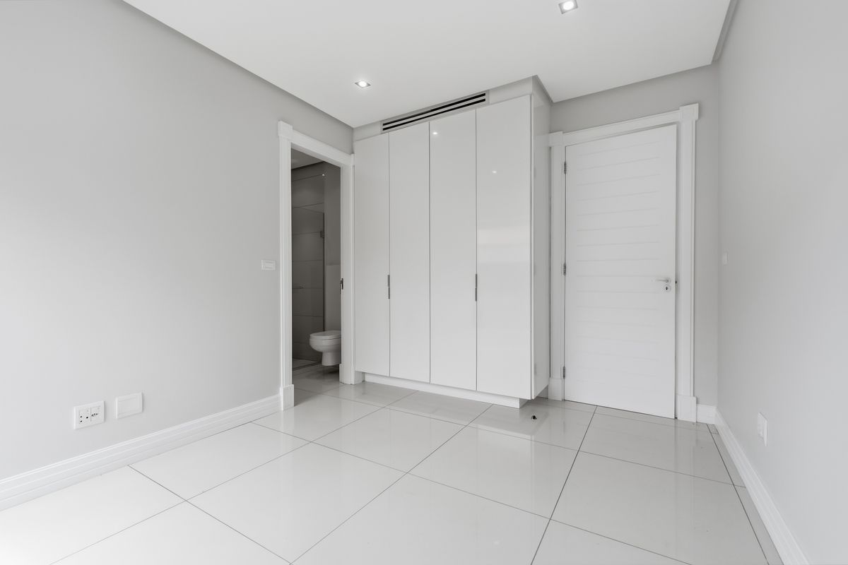 A bright, modern bedroom featuring high-gloss built-in cupboards, polished tiled flooring, and recessed lighting, with an adjacent en-suite bathroom visible.