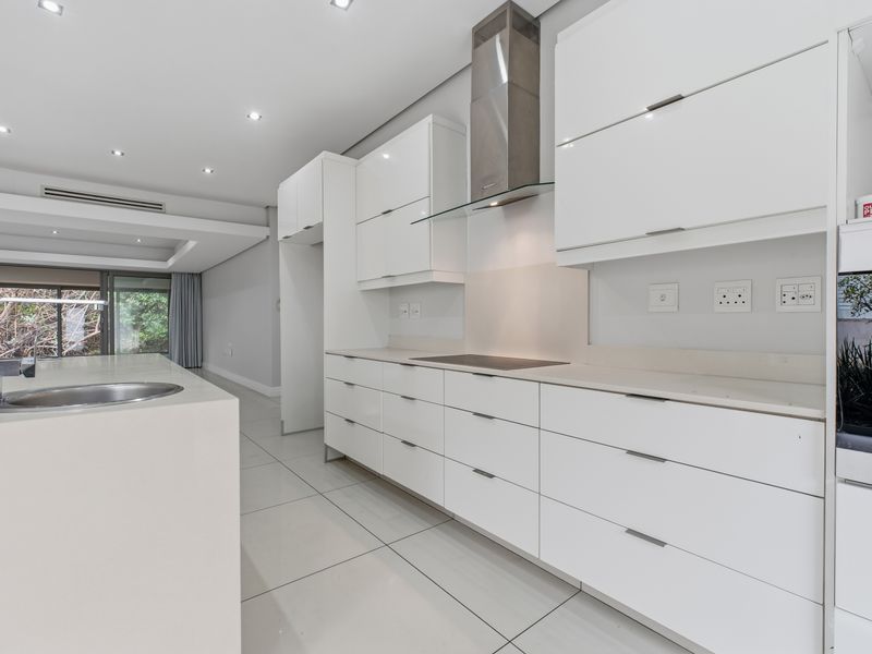 Modern, minimalist kitchen featuring sleek white cabinetry, a central island with an integrated sink, built-in oven, and high-quality tiled flooring.