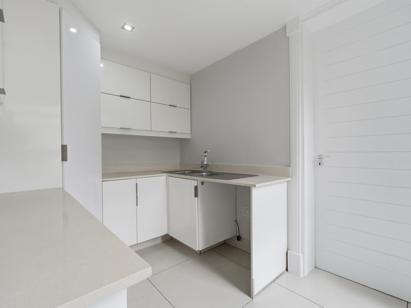 A clean, modern scullery featuring white built-in cabinetry, stone countertops, a stainless steel sink, and neutral tiled flooring.