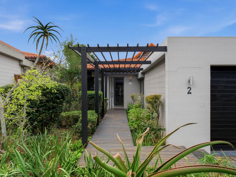 Front entrance of the townhouse featuring a timber decking walkway, black metal pergola, and a private garage entrance surrounded by landscaped garden beds.