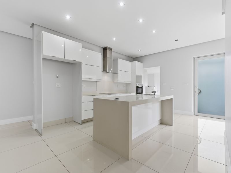 Modern, minimalist kitchen featuring a central island, high-gloss cabinetry, built-in oven, extractor fan, and sleek tiled flooring in a bright, open space.