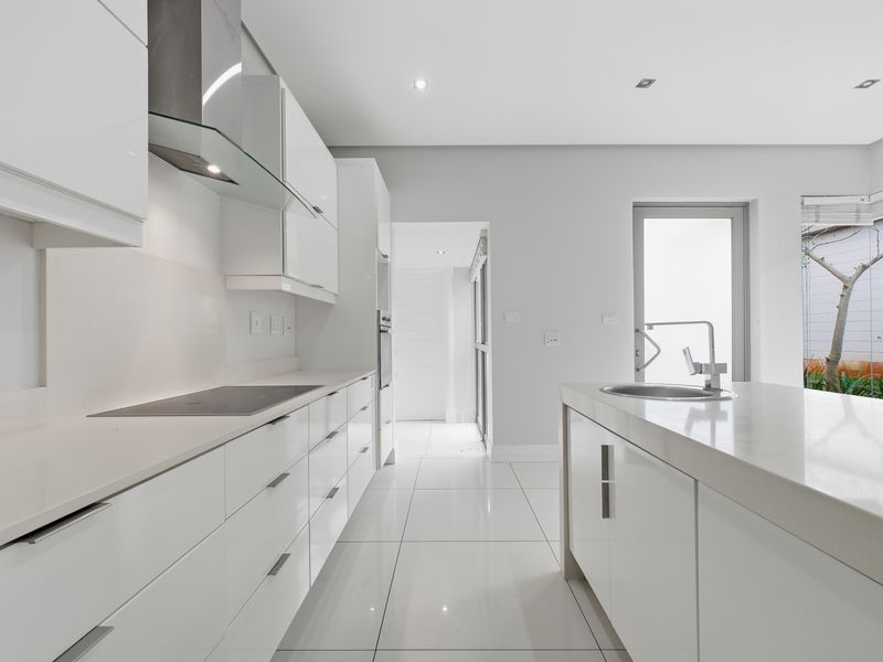 Modern, minimalist kitchen featuring white gloss cabinetry, stone countertops, a central island with sink, and high-end built-in appliances in a bright, open space.