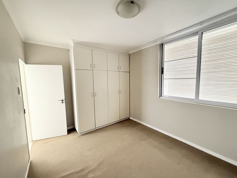 A clean, empty bedroom featuring neutral walls, carpeted flooring, and ample built-in cupboard storage with natural light from a large window.