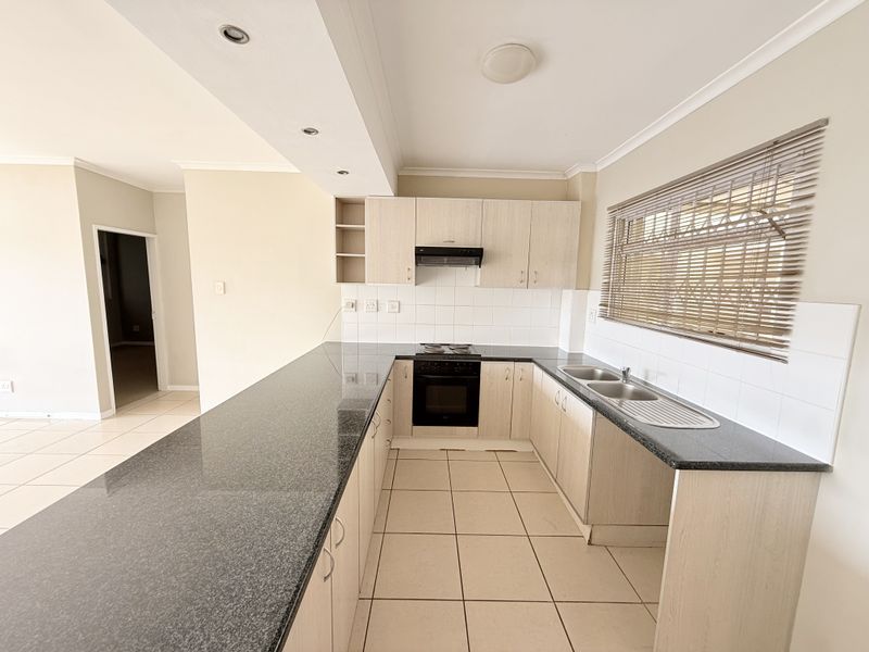 A clean, functional kitchen featuring granite countertops, built-in oven and hob, double basin sink, and ample cabinetry with neutral tiled flooring.