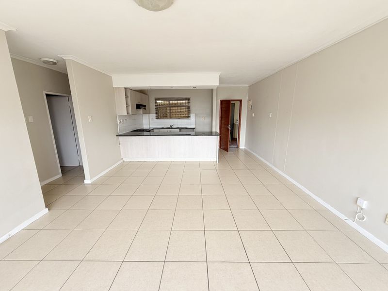 An open-plan living and kitchen area featuring neutral tiled flooring, built-in kitchen cabinetry with a dark countertop, and a functional layout.