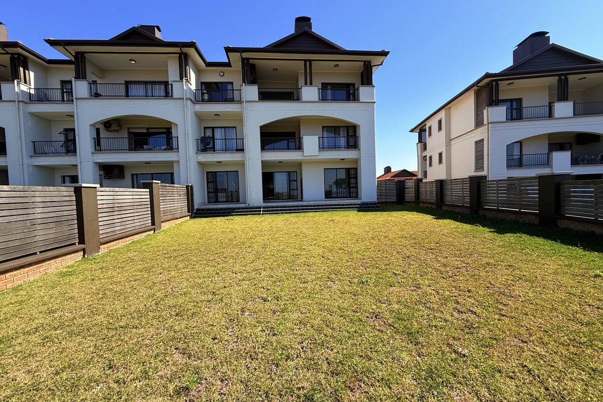A spacious rear garden area featuring a well-maintained lawn, enclosed by timber slat fencing, with views of the apartment building's rear balconies and patio.