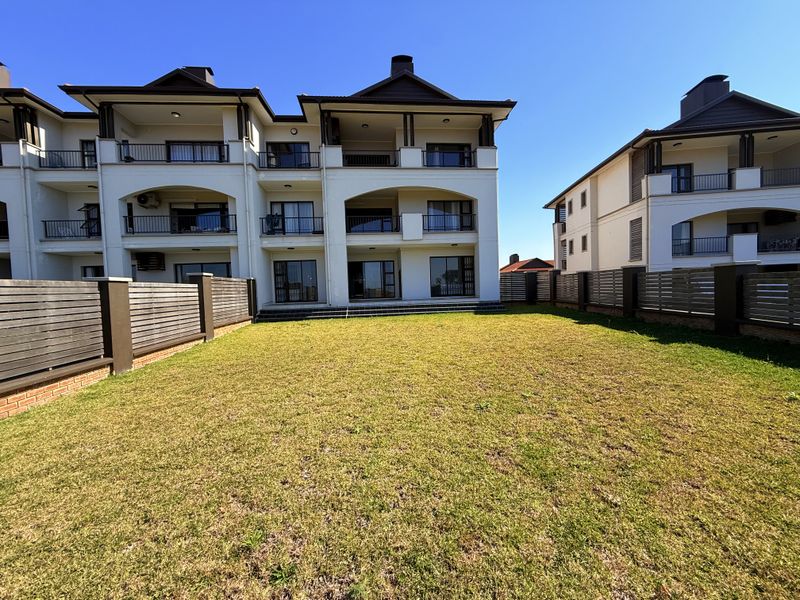 A spacious rear garden area featuring a well-maintained lawn, enclosed by timber slat fencing, with views of the apartment building's rear balconies and patio.