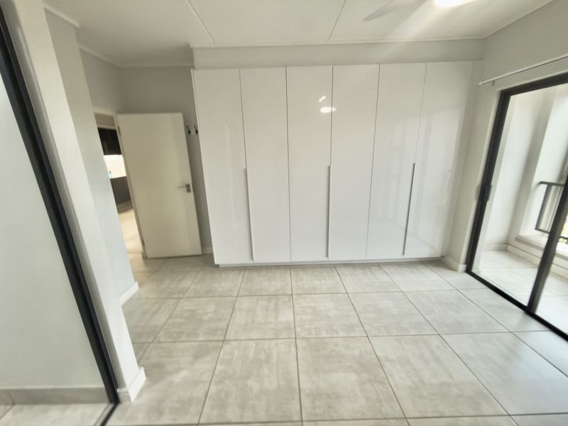 A bright, empty bedroom featuring modern white built-in cupboards, neutral tiled flooring, and a sliding glass door leading to an outdoor area.