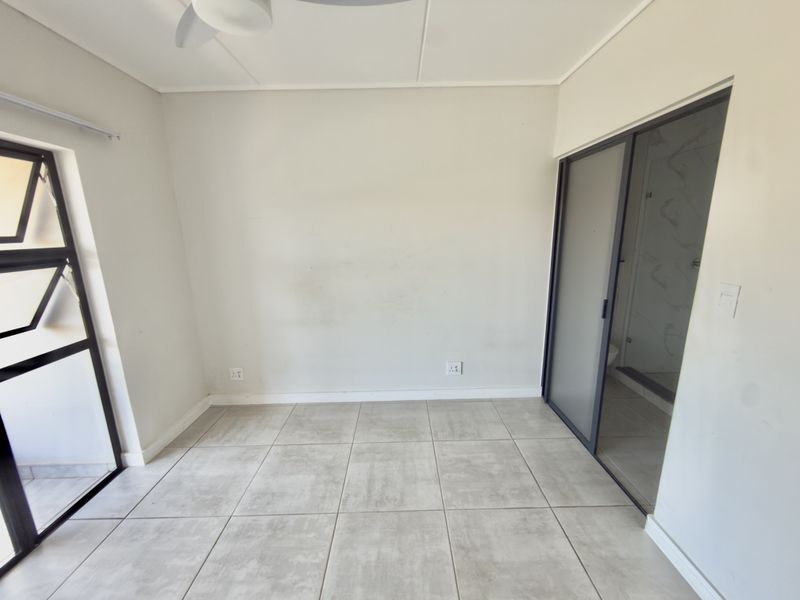 A bright, empty bedroom featuring neutral tiled flooring, a ceiling fan, and an en-suite bathroom accessible via a sliding glass door.
