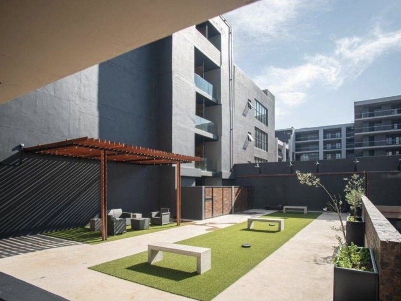 A modern communal outdoor courtyard area featuring a wooden pergola, artificial lawn, and concrete seating within an apartment complex.