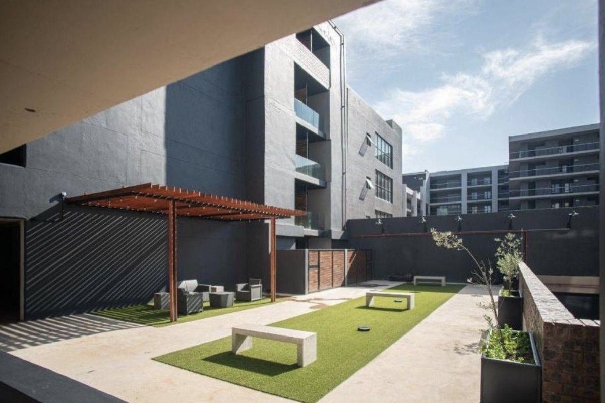 A modern communal outdoor courtyard area featuring a wooden pergola, artificial lawn, and concrete seating within an apartment complex.
