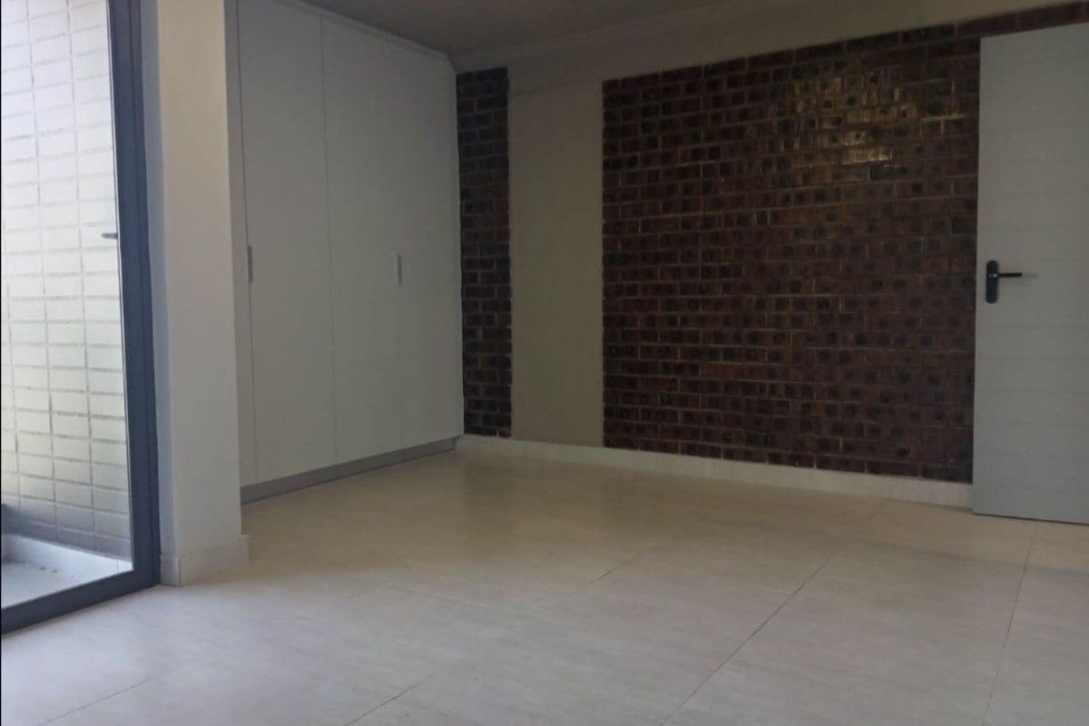 An empty bedroom featuring modern tiled flooring, built-in cupboards, and a stylish exposed brick feature wall with access to an outdoor area.