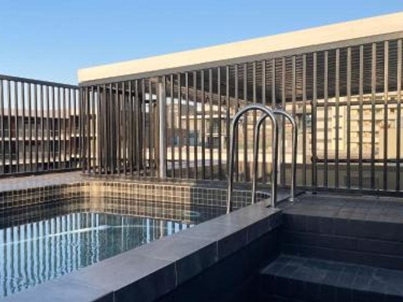 A rooftop swimming pool area featuring a tiled pool, metal safety fencing, and a covered patio structure.
