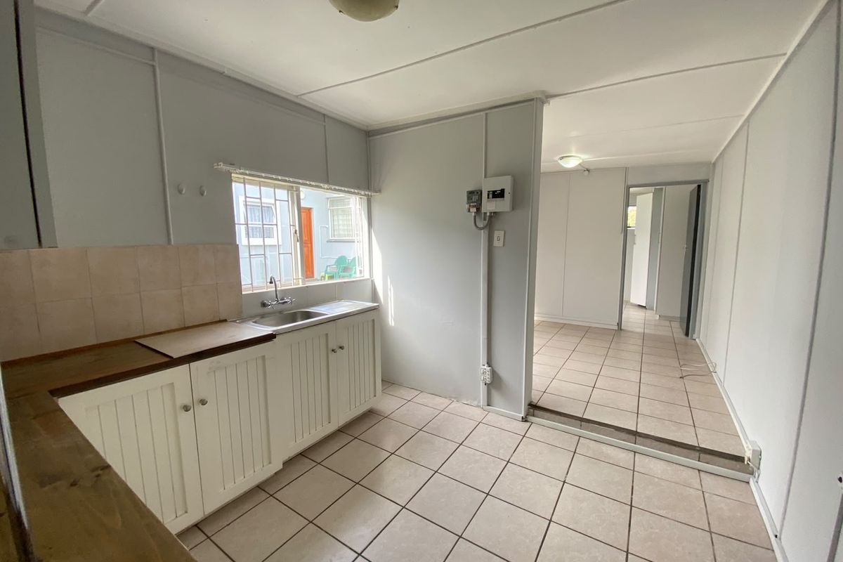 A functional, empty kitchen area featuring tiled flooring, basic cabinetry, and a stainless steel sink, leading into an adjacent living space.
