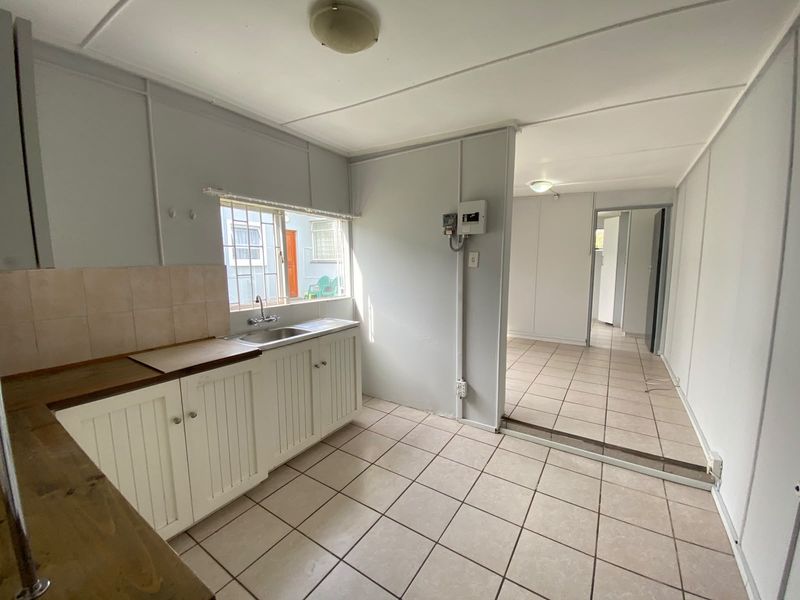 A functional, empty kitchen area featuring tiled flooring, basic cabinetry, and a stainless steel sink, leading into an adjacent living space.