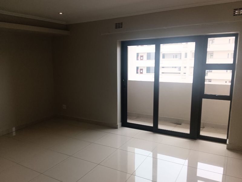 An empty, well-lit living area featuring neutral tiled flooring and a large sliding glass door providing access to an outdoor balcony.