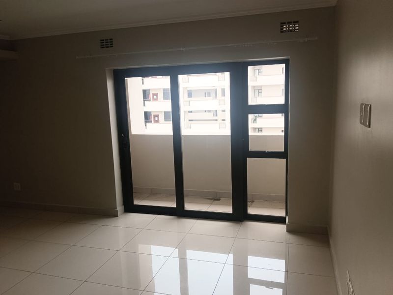 An empty, well-maintained living area featuring neutral tiled flooring and a large sliding glass door providing access to an outdoor balcony.