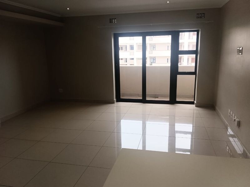 A spacious, empty living area featuring neutral tiled flooring, recessed lighting, and a large sliding glass door providing access to an outdoor balcony.