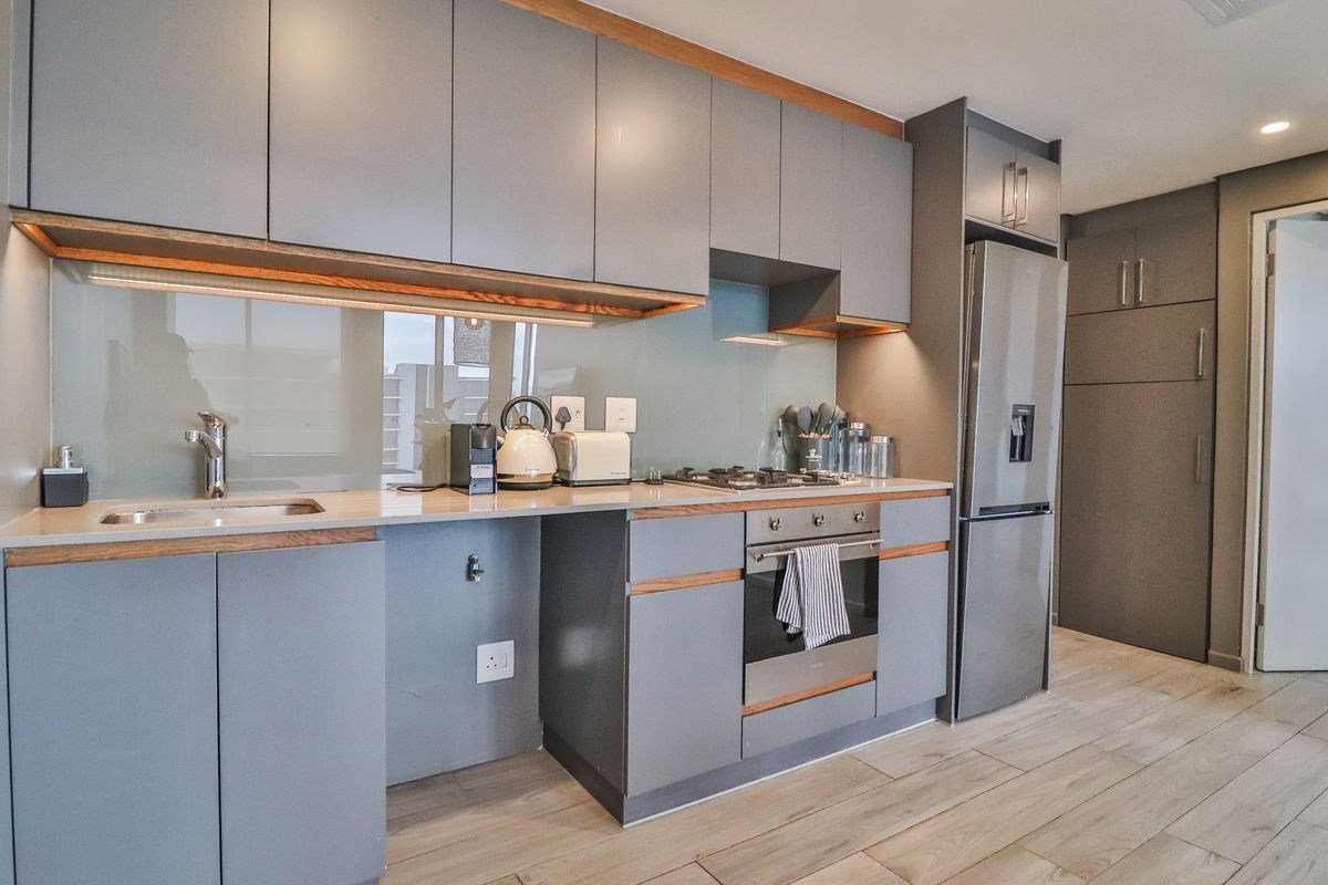 Modern kitchen featuring sleek grey cabinetry, stone countertops, glass splashback, integrated appliances, and high-quality wood-look tile flooring with warm under-cabinet lighting.