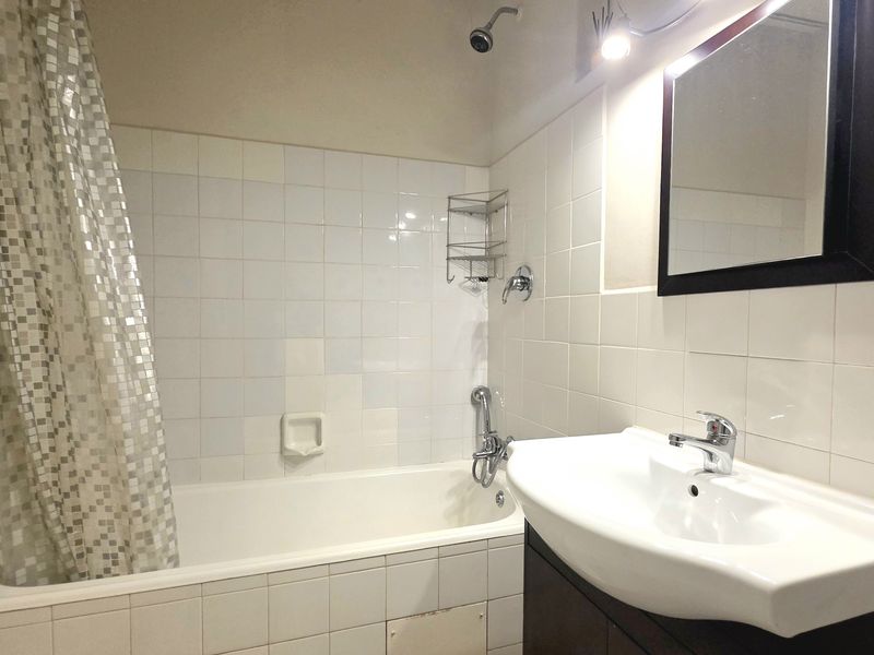 A functional bathroom featuring a bathtub with shower mixer, wall-mounted vanity basin with cabinet, and white tiled walls.