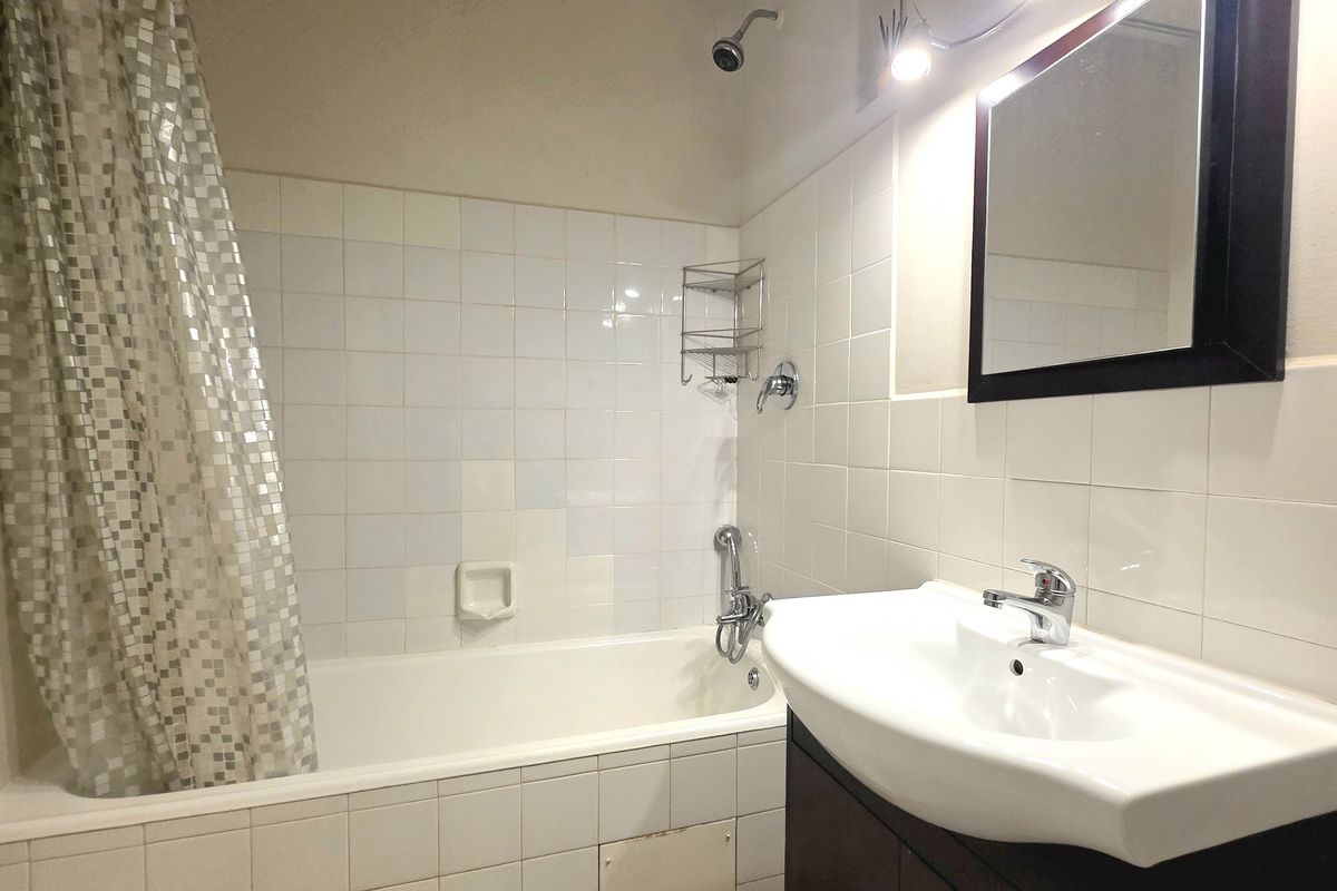 A functional bathroom featuring a bathtub with shower mixer, wall-mounted vanity basin with cabinet, and white tiled walls.