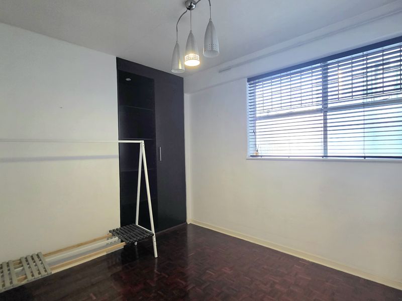 A bright, empty bedroom featuring dark parquet flooring, a built-in wardrobe, and venetian blinds, offering a clean canvas for personalization.
