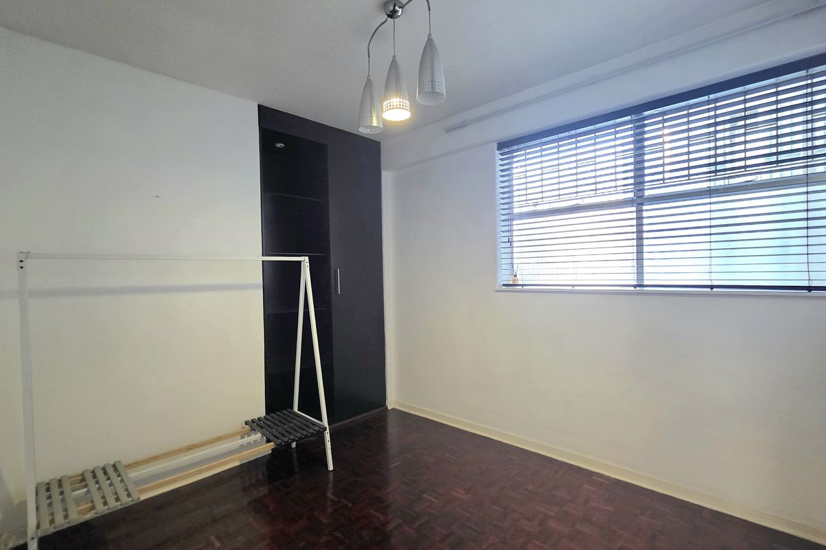 A bright, empty bedroom featuring dark parquet flooring, a built-in wardrobe, and venetian blinds, offering a clean canvas for personalization.