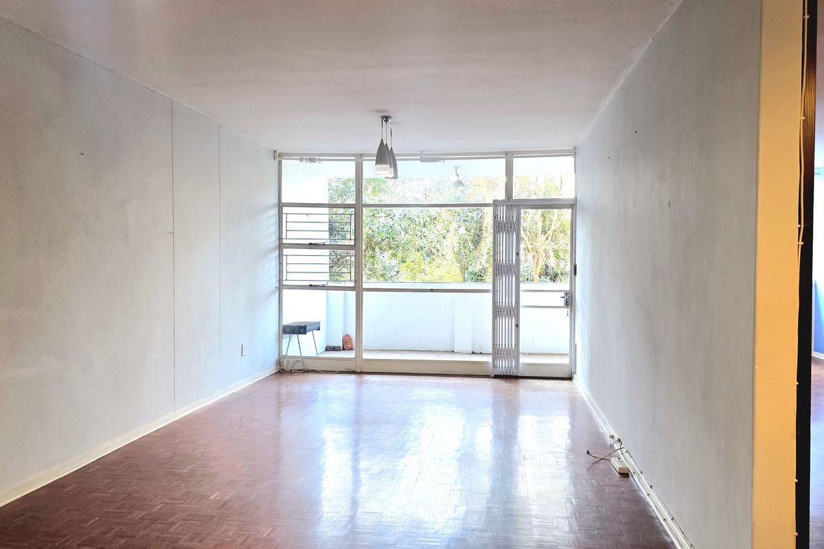 An empty, sunlit living room featuring parquet flooring, large windows, and direct access to a balcony through a sliding glass door.