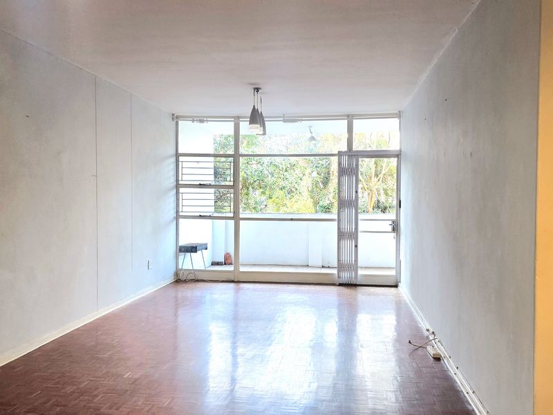 An empty, sunlit living room featuring parquet flooring, large windows, and direct access to a balcony through a sliding glass door.