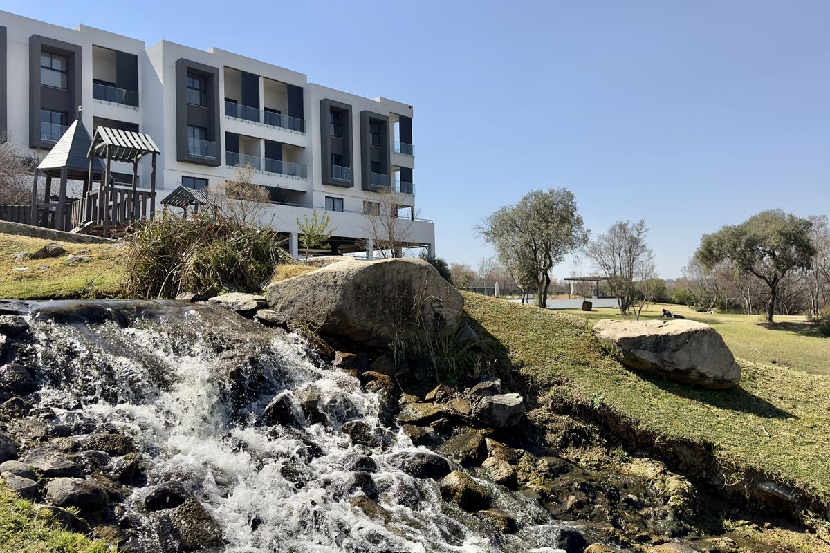 A scenic view of the apartment complex exterior featuring a landscaped garden, a natural-style water feature, and a children's playground area.
