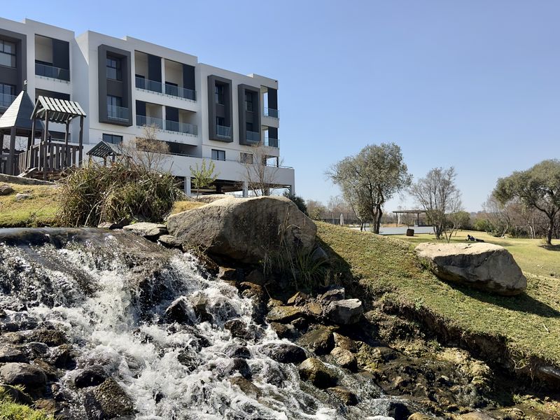 A scenic view of the apartment complex exterior featuring a landscaped garden, a natural-style water feature, and a children's playground area.