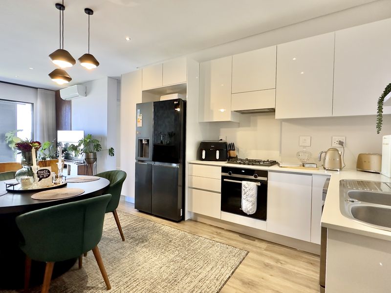Modern open-plan kitchen and dining area featuring sleek white cabinetry, integrated appliances, laminate flooring, and contemporary lighting fixtures.