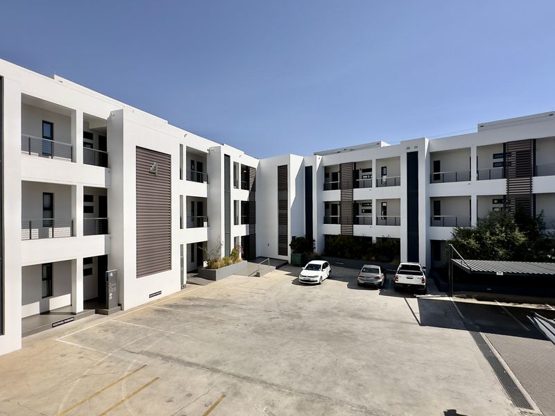 A modern apartment complex exterior featuring a paved courtyard, covered parking bays, and multi-level balconies with contemporary architectural design.