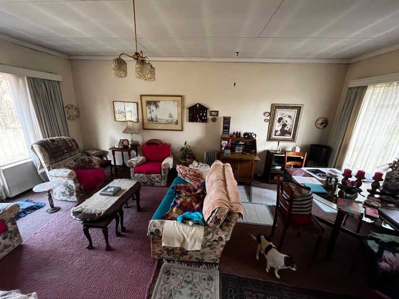 A spacious living room featuring carpeted flooring and large windows with curtain tracks, offering a dated interior ready for modernization.