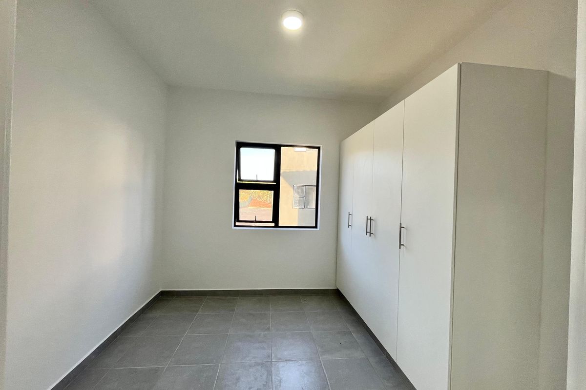 A clean, empty bedroom featuring modern grey floor tiles, white walls, and ample built-in cupboard storage with a window providing natural light.