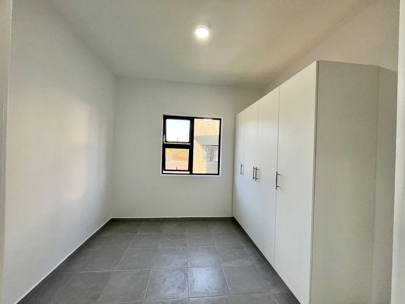 A clean, empty bedroom featuring modern grey floor tiles, white walls, and ample built-in cupboard storage with a window providing natural light.