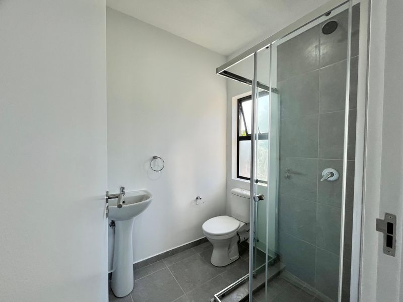 A clean, modern bathroom featuring a pedestal basin, toilet, and a glass-enclosed shower with grey tiled finishes.