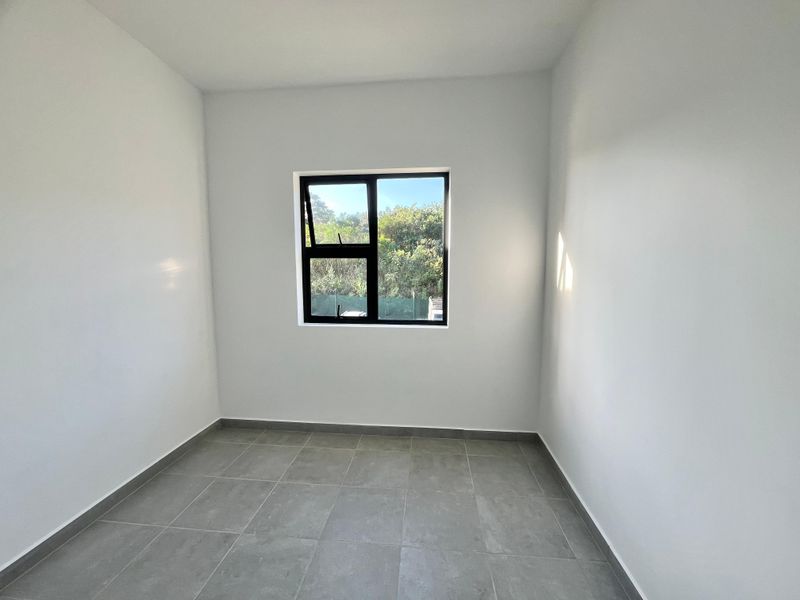 A clean, empty bedroom featuring neutral walls, modern grey floor tiles, and a black aluminium window frame with natural light.