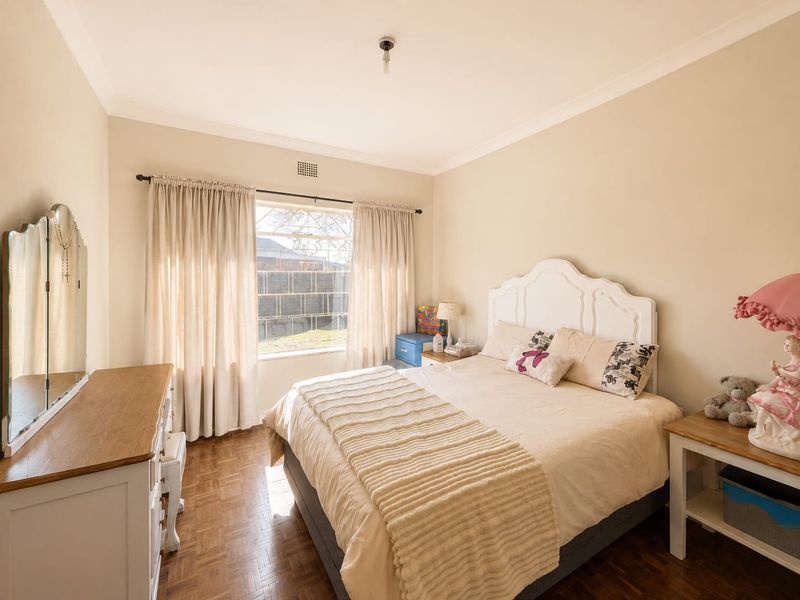 A well-lit bedroom featuring classic parquet flooring, neutral wall tones, and a large window with curtain tracks, providing a comfortable and functional living space.