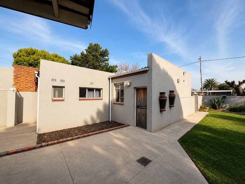 A well-maintained rear exterior view featuring a concrete patio, manicured lawn, and a stucco-finished building facade with timber door and casement windows.