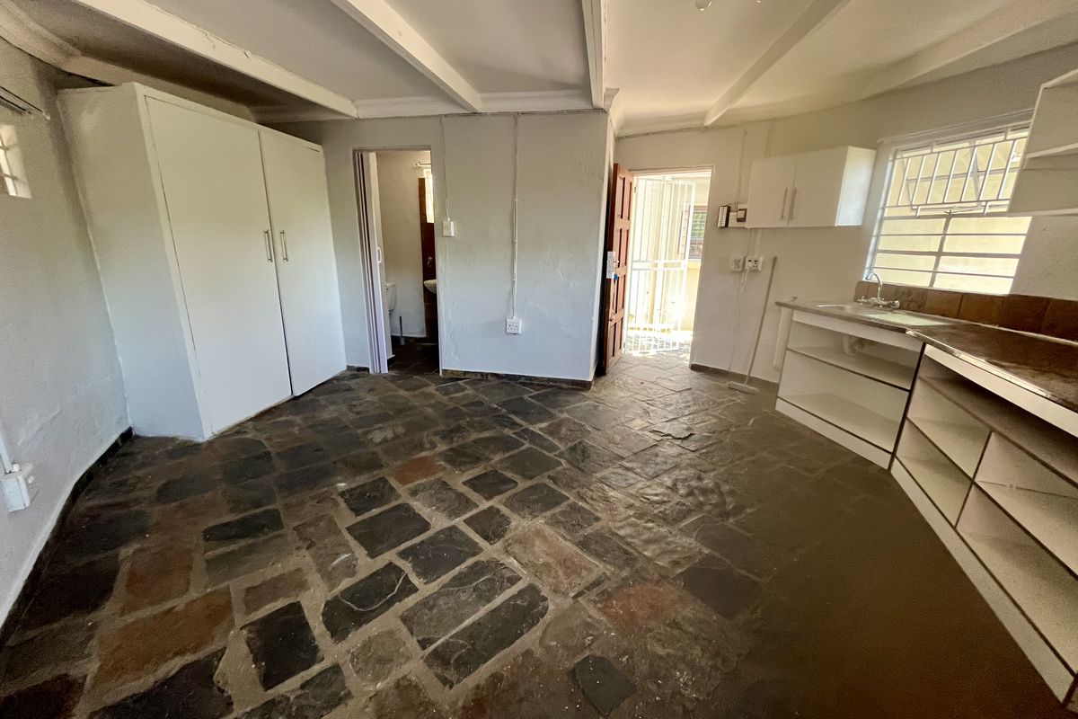 Open plan living area featuring stone tile flooring, exposed ceiling beams, built-in cupboards, and a basic kitchenette with sink and wall-mounted storage.
