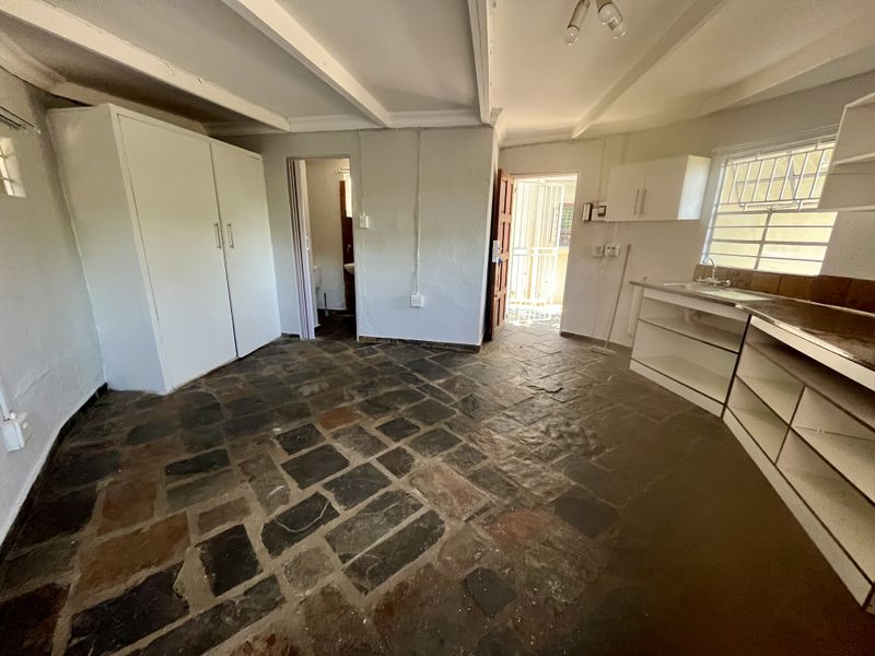 Open plan living area featuring stone tile flooring, exposed ceiling beams, built-in cupboards, and a basic kitchenette with sink and wall-mounted storage.
