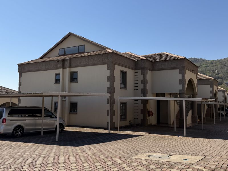 Exterior view of a multi-story apartment complex featuring a paved driveway, covered carports, and a tiled roof with stone-accented facade details.
