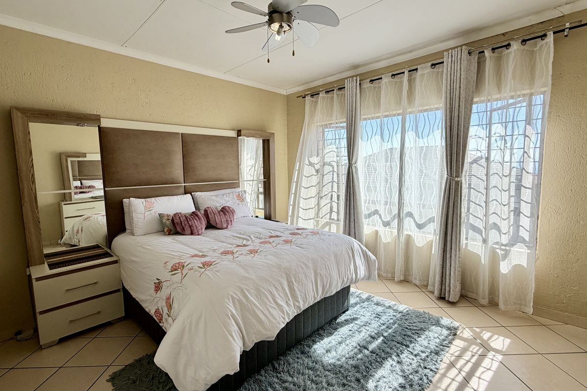 A well-lit bedroom featuring tiled flooring, a ceiling fan, and large windows with security bars, offering a comfortable and functional living space.