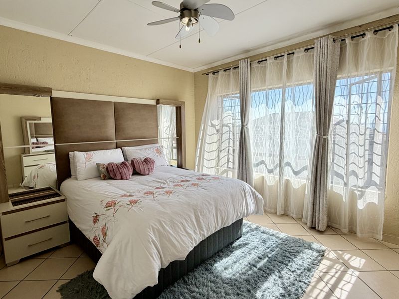A well-lit bedroom featuring tiled flooring, a ceiling fan, and large windows with security bars, offering a comfortable and functional living space.