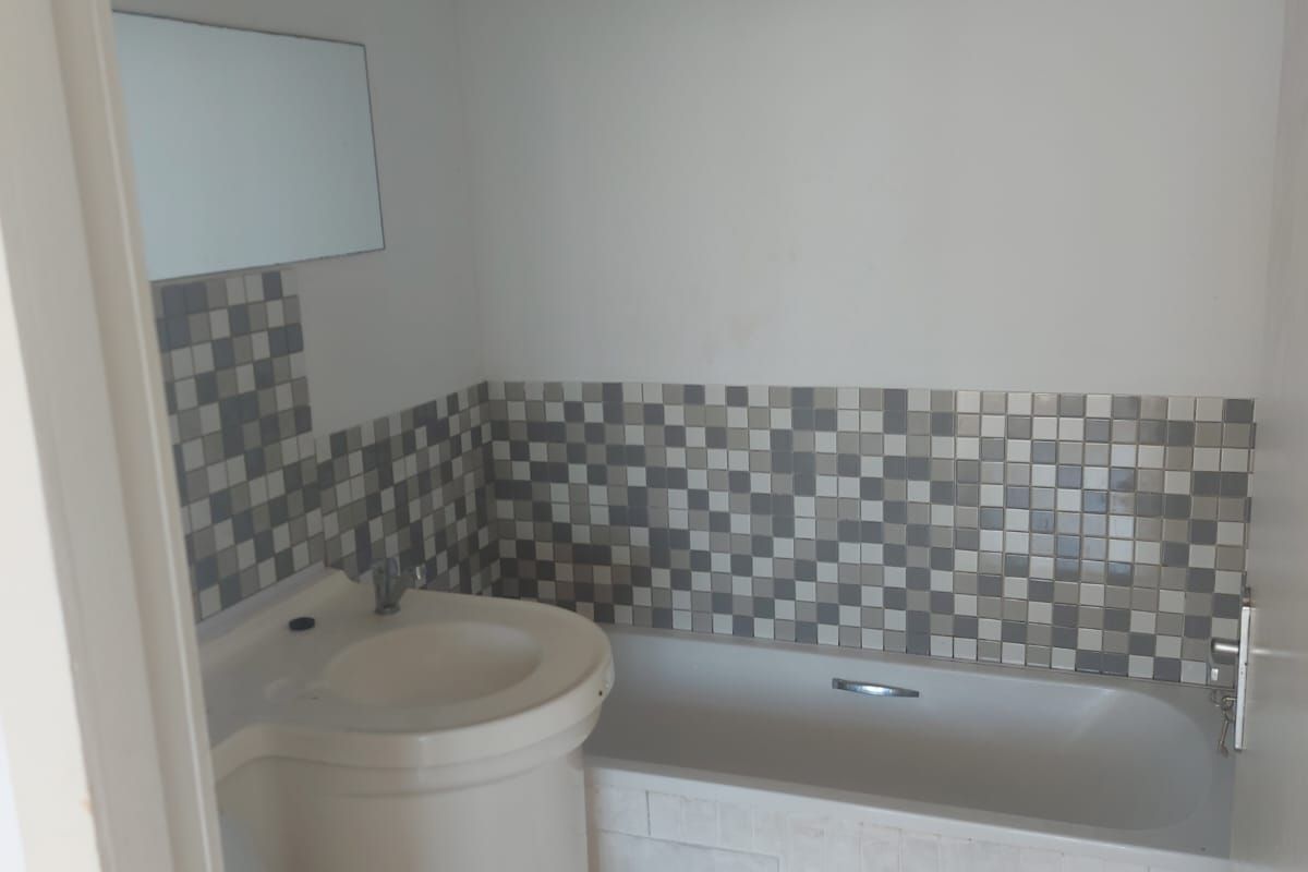 A compact bathroom featuring a pedestal basin, bathtub with tiled splashback, and tiled flooring. The space is functional but requires cosmetic updates.