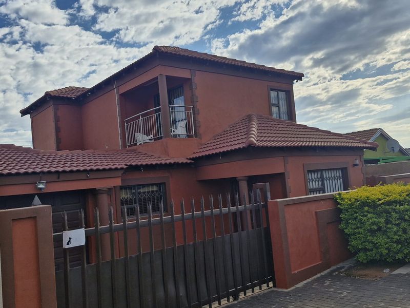 A two-story residential house featuring a terracotta-colored facade, tiled roof, upper-level balcony, and a secure gated entrance with a paved driveway.