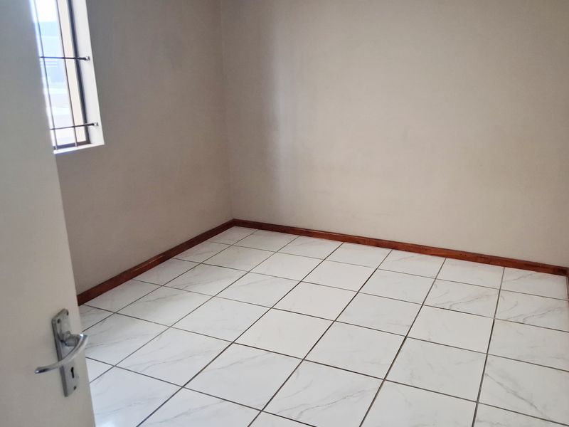 A clean, empty bedroom featuring white marble-look tiled flooring, neutral walls, and a window fitted with security bars.