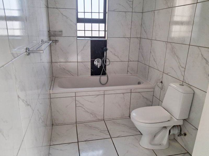 A clean, modern bathroom featuring white marble-look wall and floor tiles, a built-in bathtub with shower mixer, and a standard toilet.