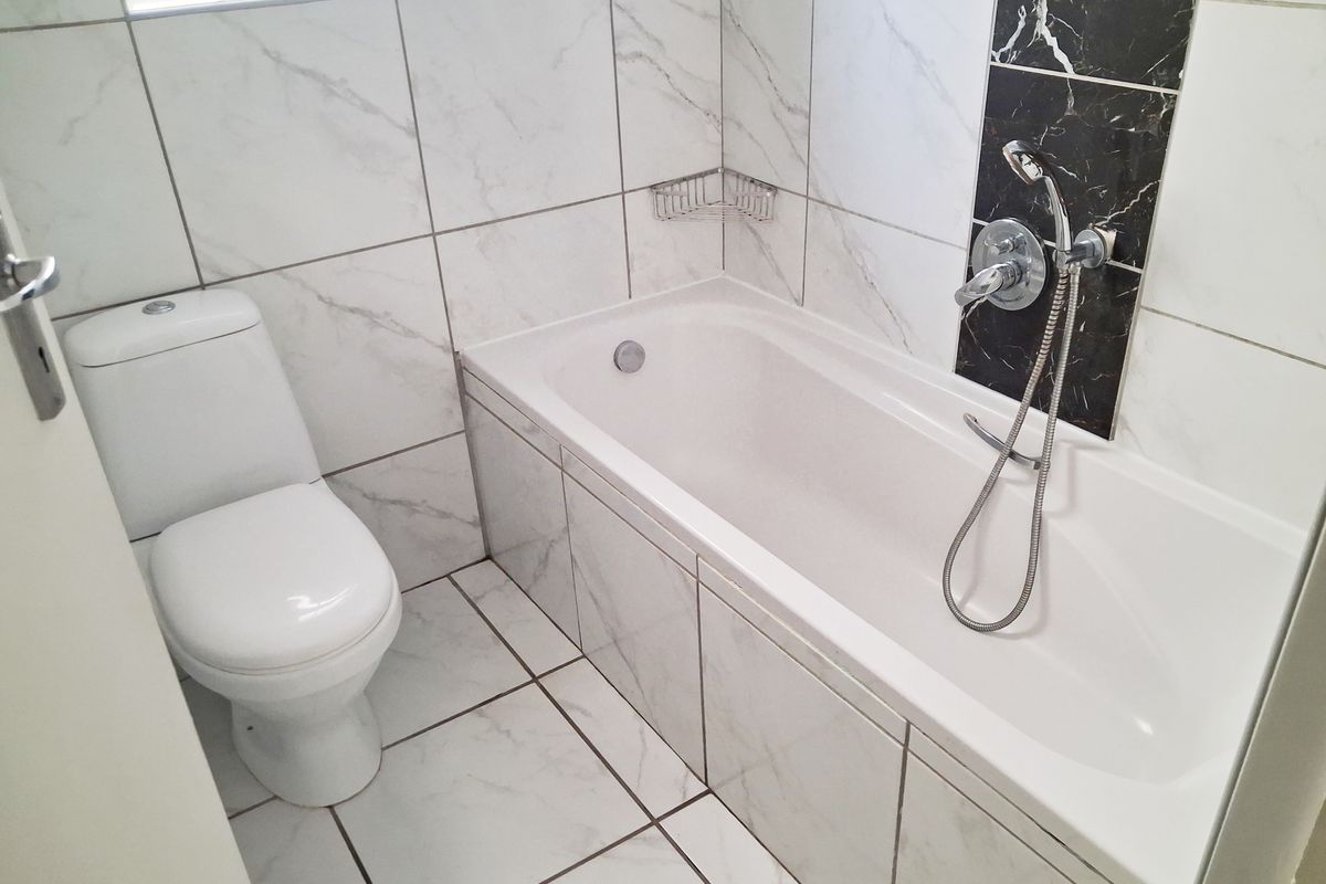 A clean, modern bathroom featuring white marble-look wall and floor tiles, a built-in bathtub with a shower mixer, and a standard toilet.